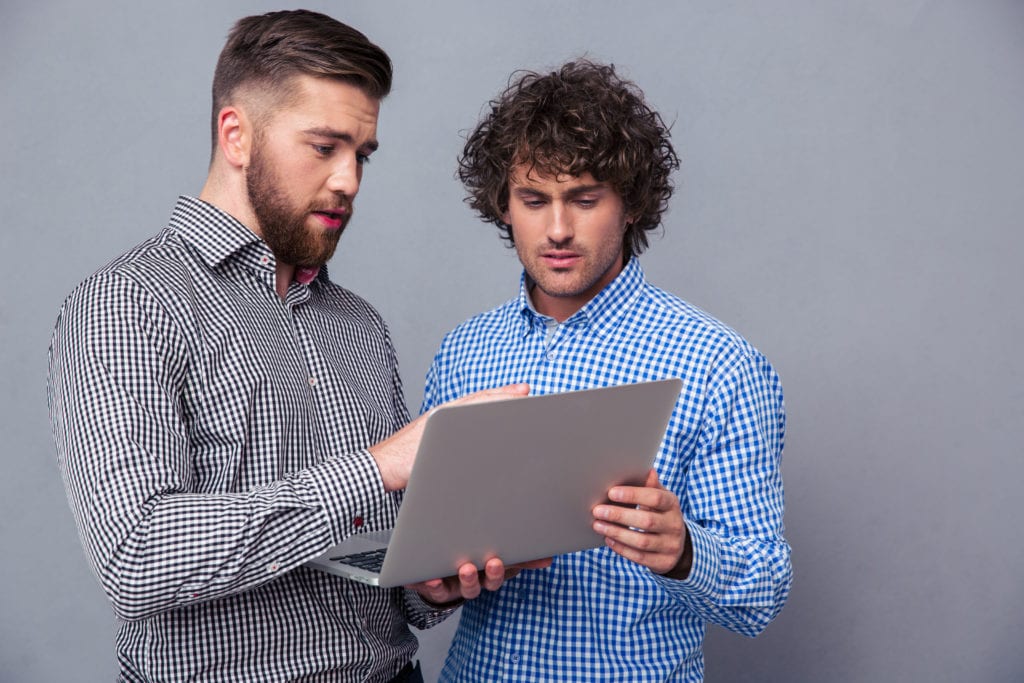 two handsome men using a laptop together, one showing the other something interesting, over a gray background