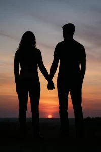 Silhouette of a couple holding hands at sunset, standing close together against a colorful sky with orange, pink, and purple hues.